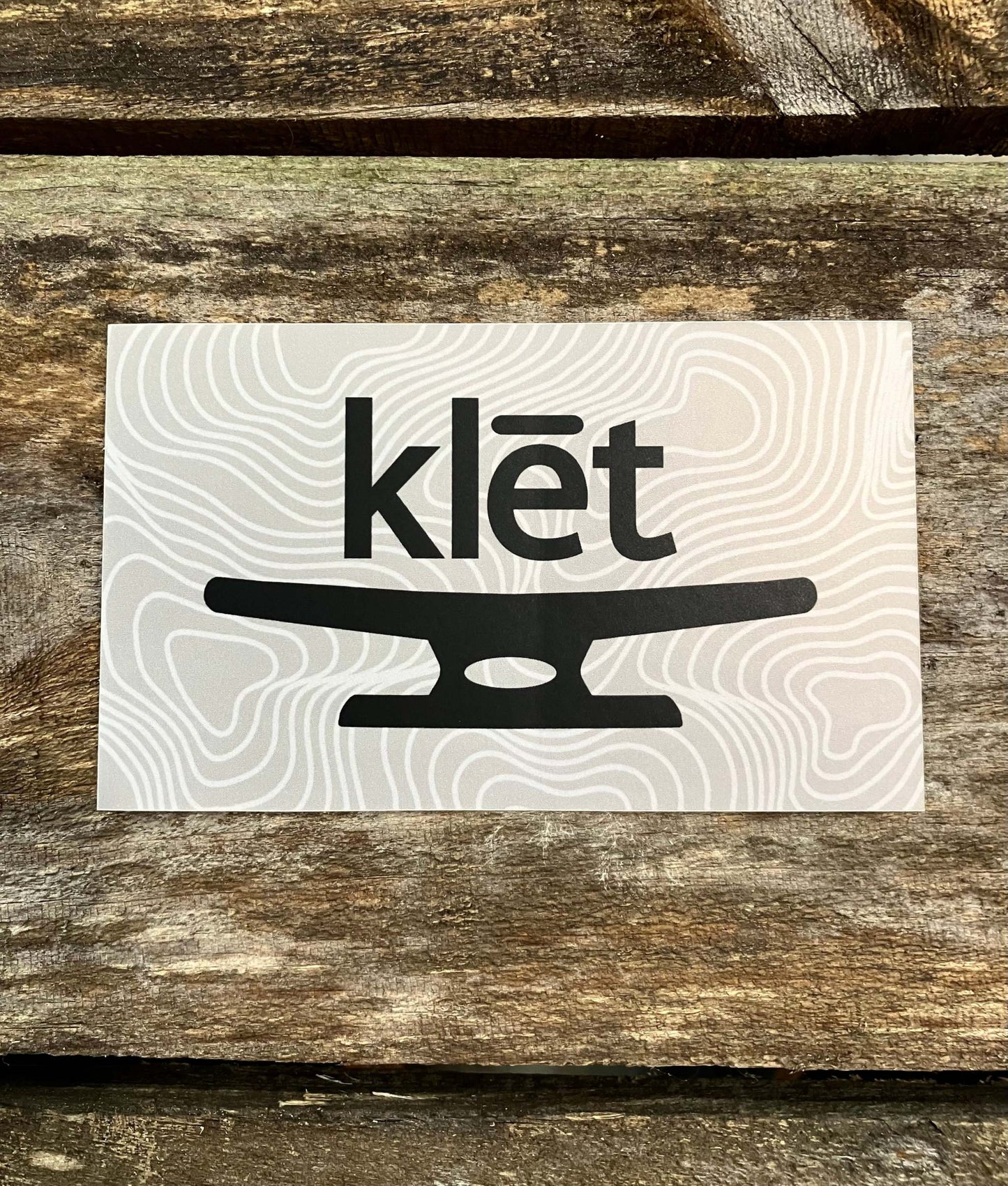 Klēt Open Sea 5-inch sticker with black logo on white topographic background placed on wooden surface