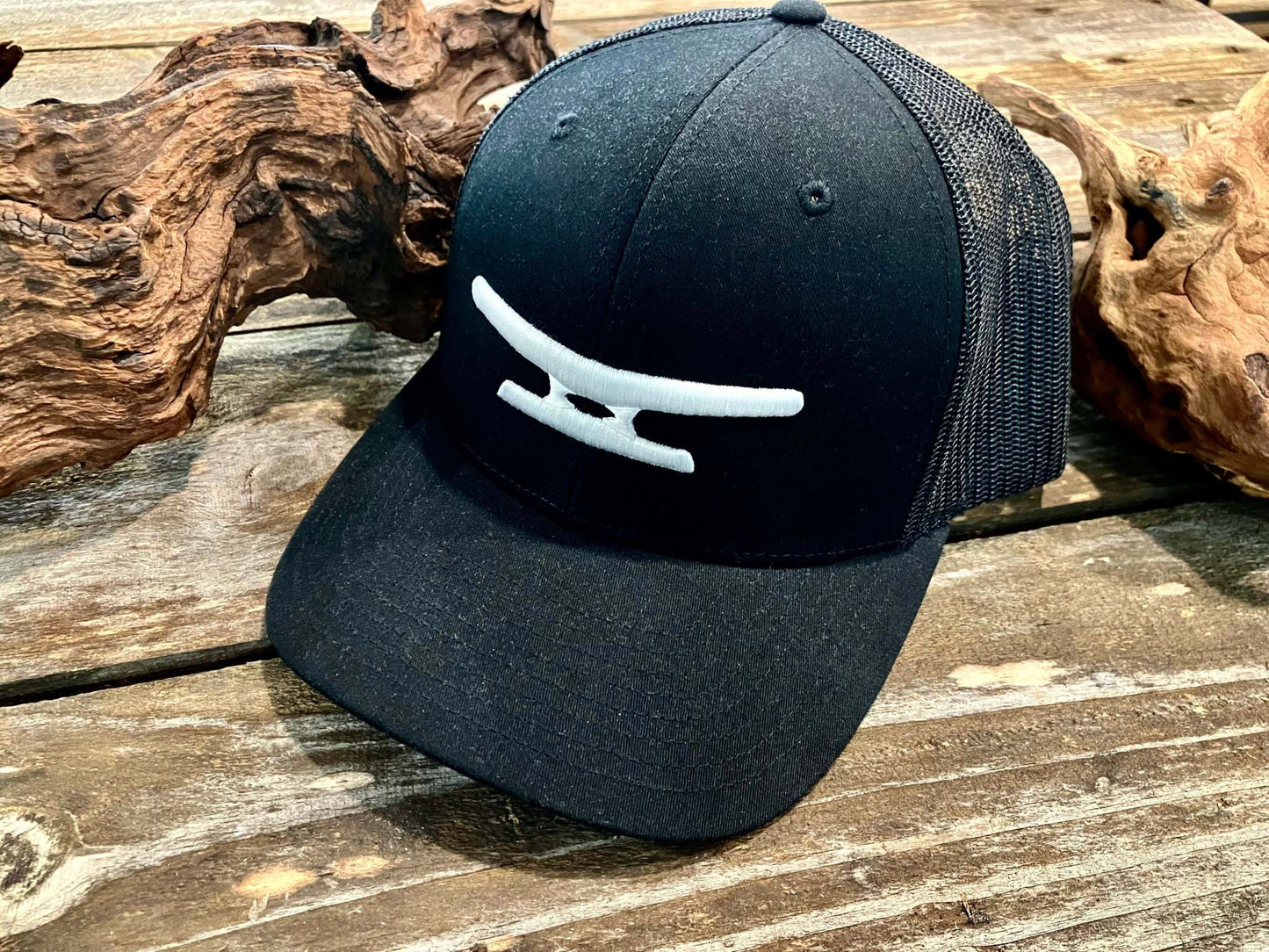 Black Klēt trucker hat with white puff embroidered monogram logo on front panel placed on rustic wooden surface