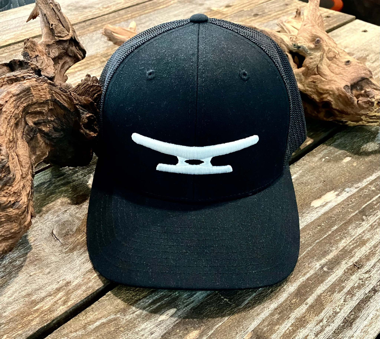 Black mesh cap with white puff embroidered Klēt monogram logo on front panel placed on wooden surface with driftwood pieces.
