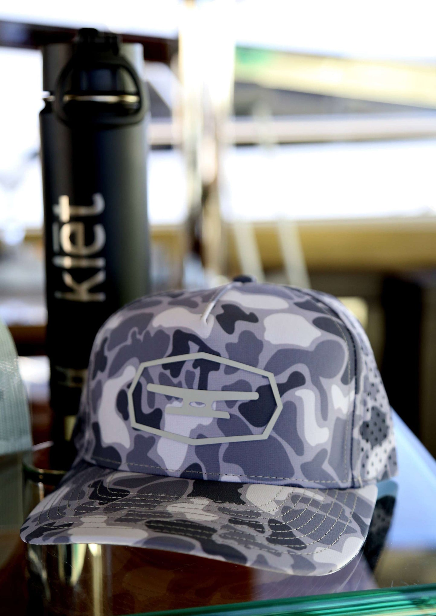Klēt Commodore Camo hat with gray camouflage pattern and branded logo, displayed alongside a black Klēt water bottle.