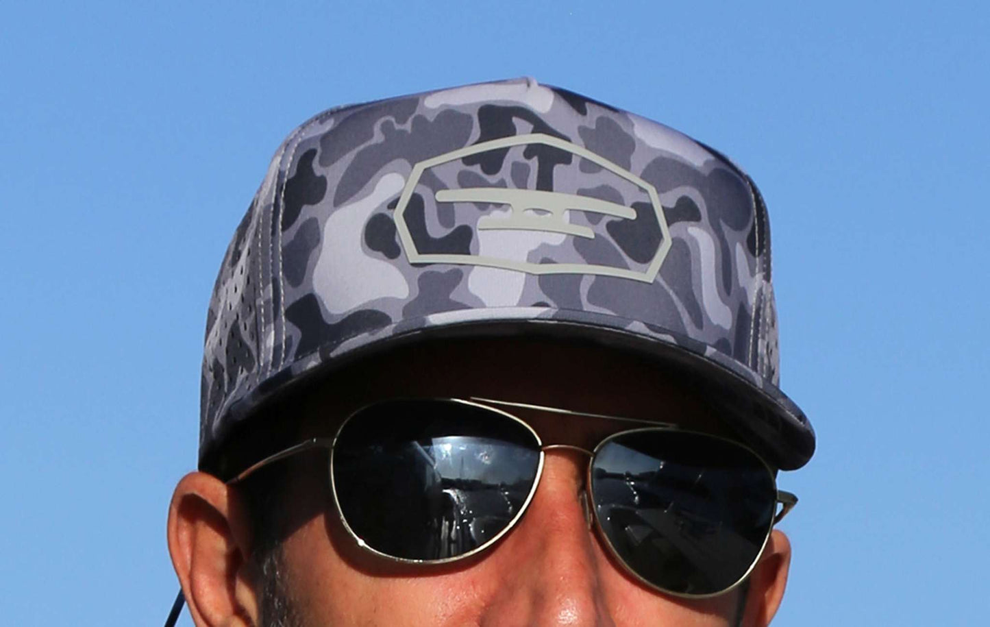 Man wearing Klēt Commodore Camo camo-patterned cap and aviator sunglasses against clear blue sky