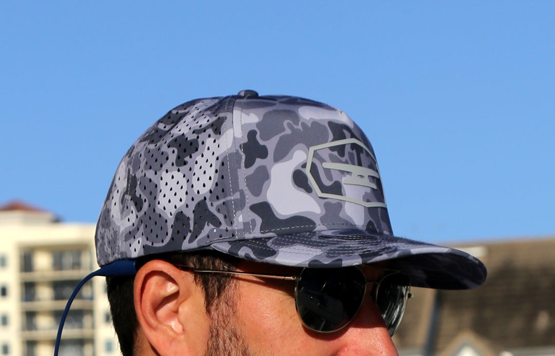 Camouflage patterned Klēt Commodore Camo cap worn by a man with sunglasses against a clear blue sky.