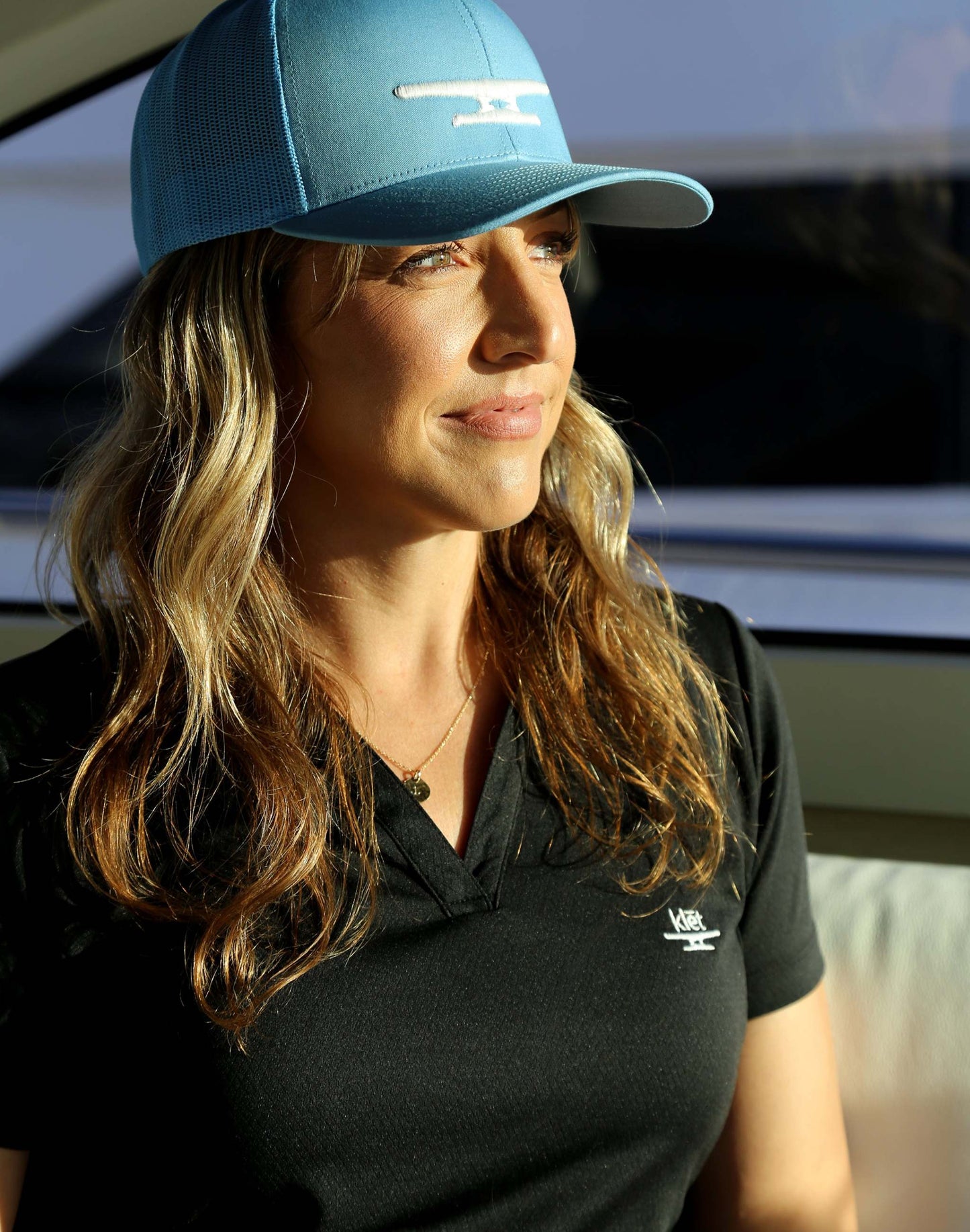 Woman wearing a black shirt with Klēt monogram puff embroidery and a blue cap, sitting in natural light